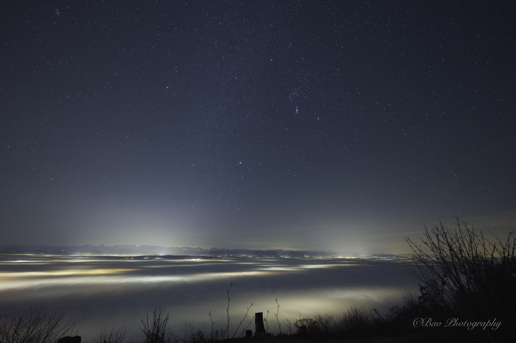 Sea of fog at night seen from Magglingen with city lights glowing beneath and a starry sky with Orion above the Alps