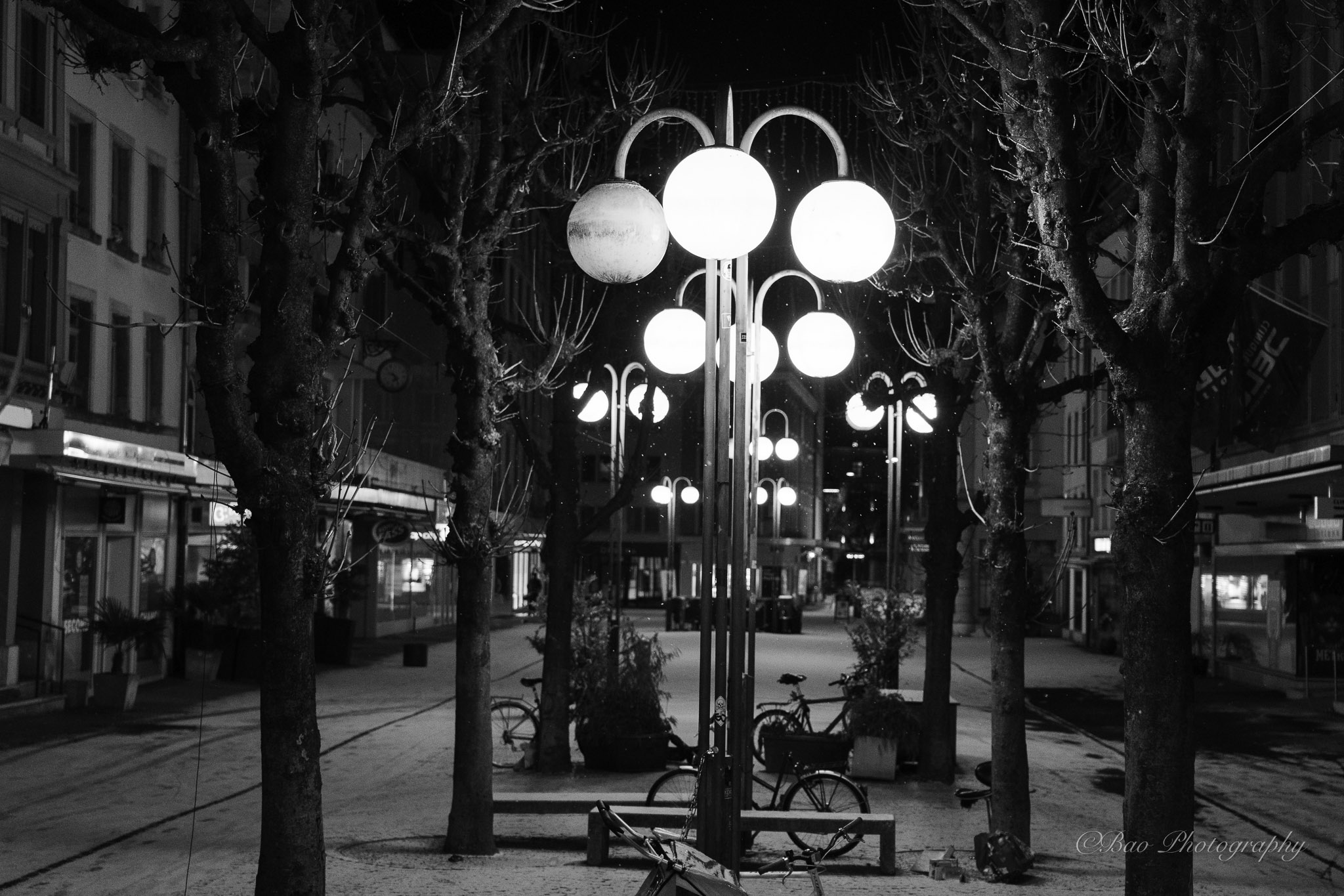 Black and white winter night scene of glowing globe street lamps near Nidaugasse in Biel on a tree-lined pedestrian street
