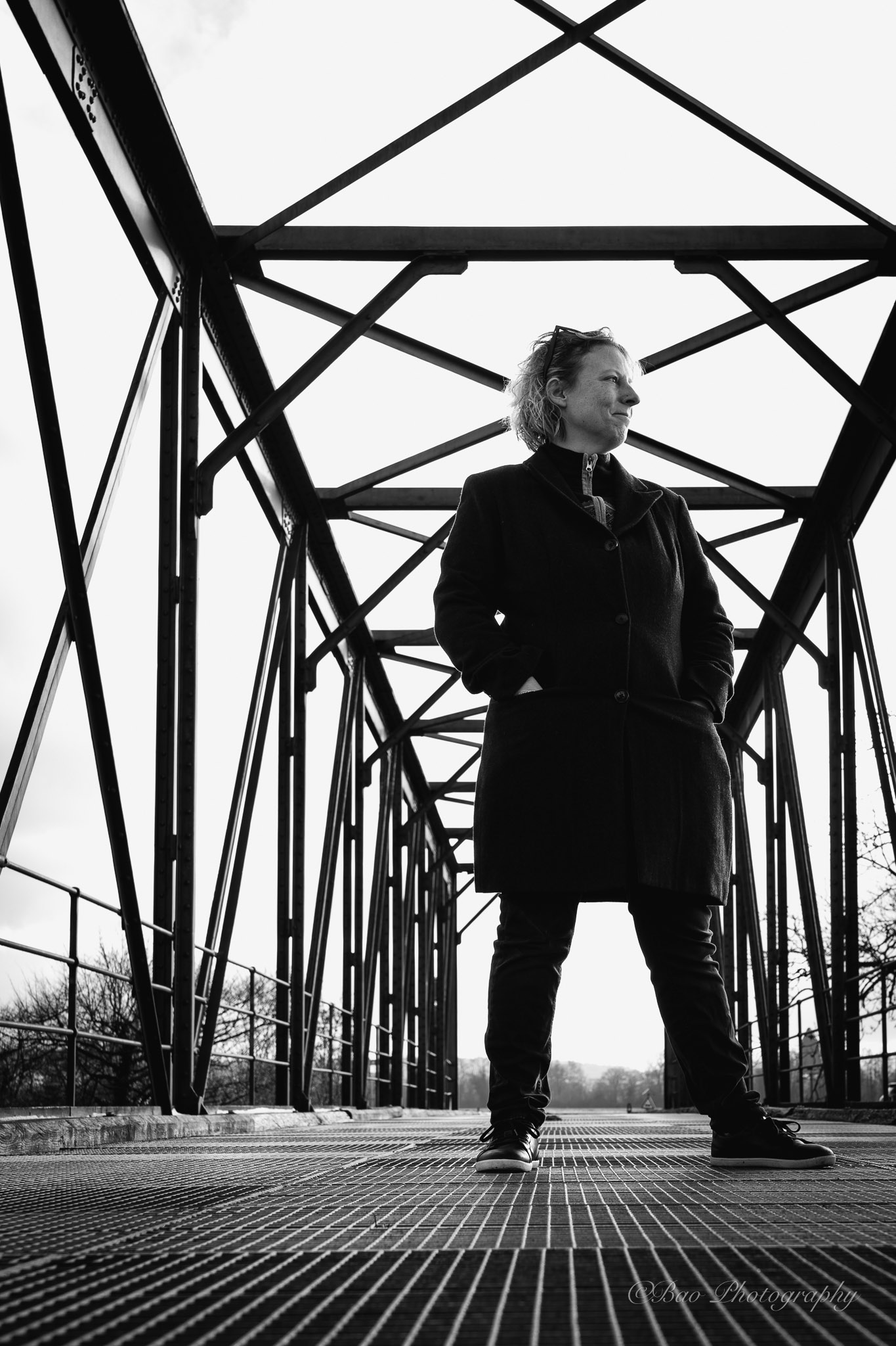 Black and white low-angle portrait of Sara standing on the Walperswilbrücke with geometric steel beams framing the composition
