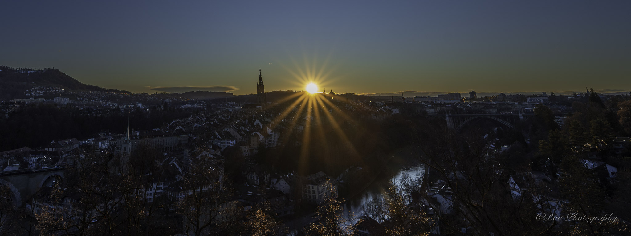 Panoramic sunset over Bern seen from the Rosengarten with the Münster cathedral spire silhouetted against a starburst sun and the Aare river below