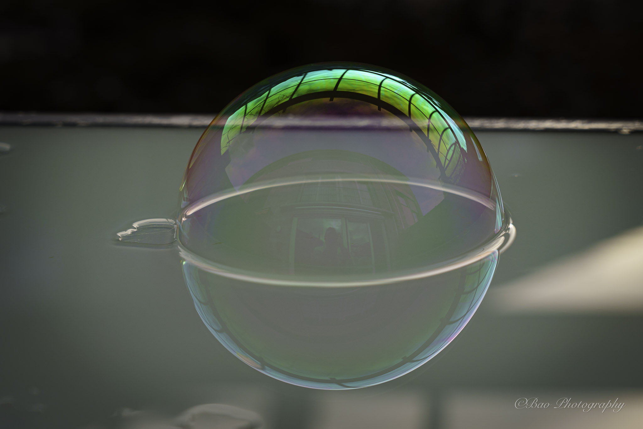 Macro close-up of a soap bubble resting on a glass surface with iridescent green and purple reflections and the photographer's silhouette visible inside