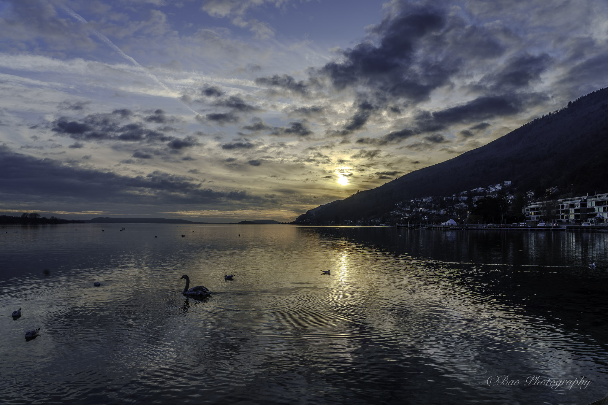 Sunset over Lake Biel with swans on the water, the sun breaking through dramatic clouds and reflecting on the calm surface, with the hillside of Magglingen and the Biel lakefront on the right