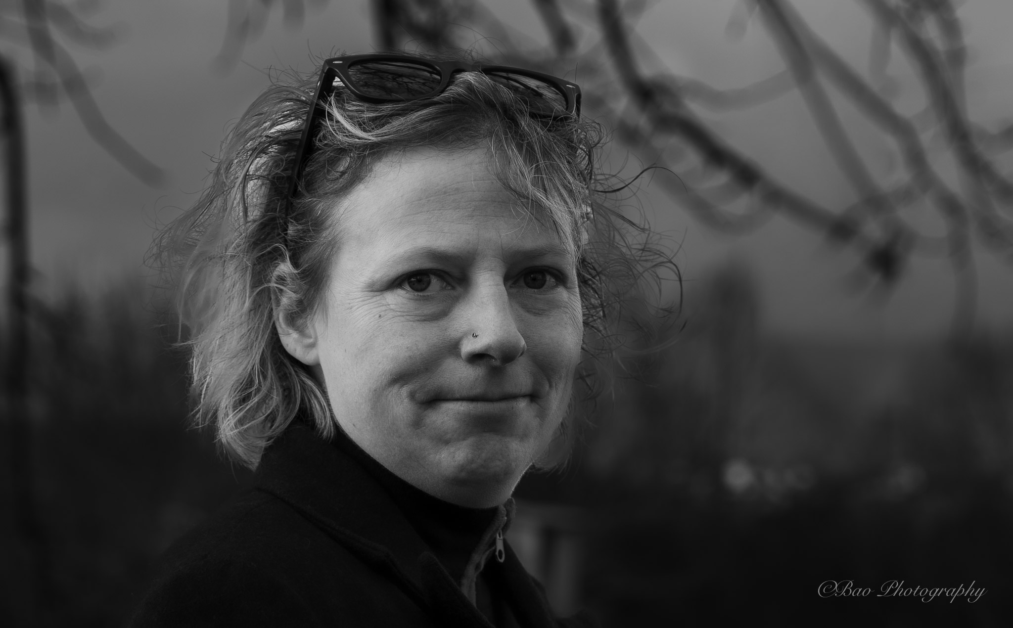 Black and white portrait of Sara with sunglasses on her head and a nose piercing, curly hair caught by the wind, with bare tree branches in the blurred background