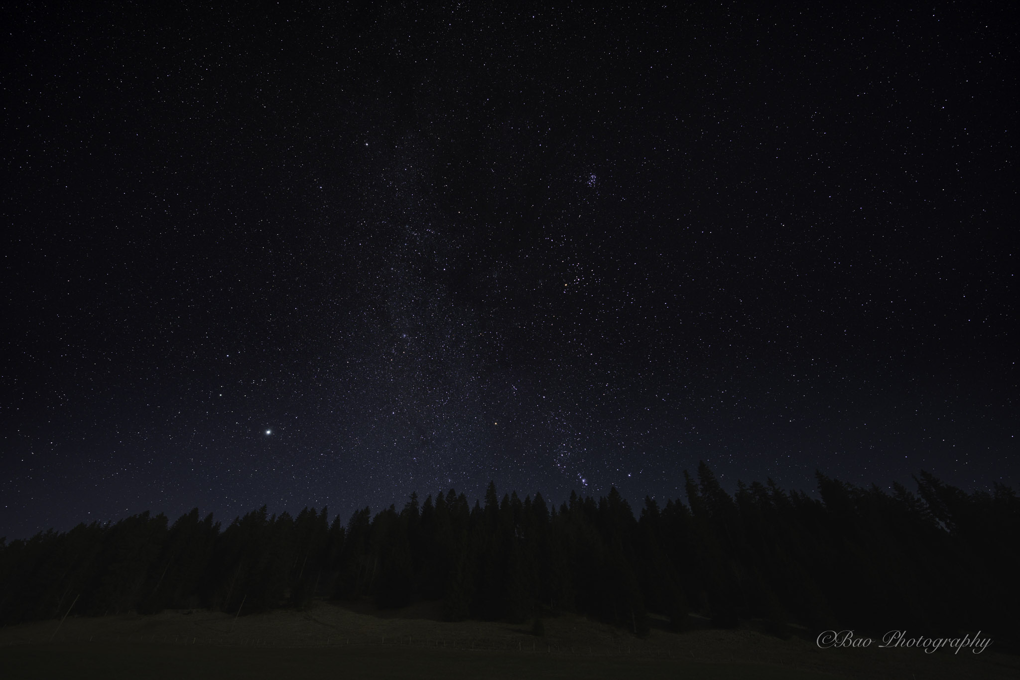 Stacked night sky photograph showing countless stars and the Milky Way above a dark conifer forest silhouette in La Brevine, with a bright planet shining on the left