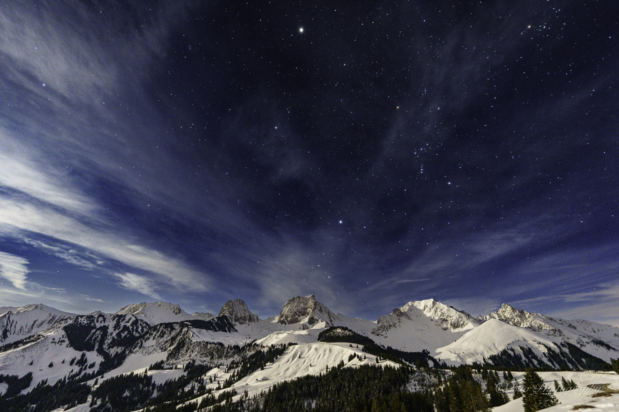 Starry night sky with wispy clouds over the snow-covered Gantrisch massif seen from Gurnigelpass, with Alpine peaks illuminated by moonlight and dark conifer forests below