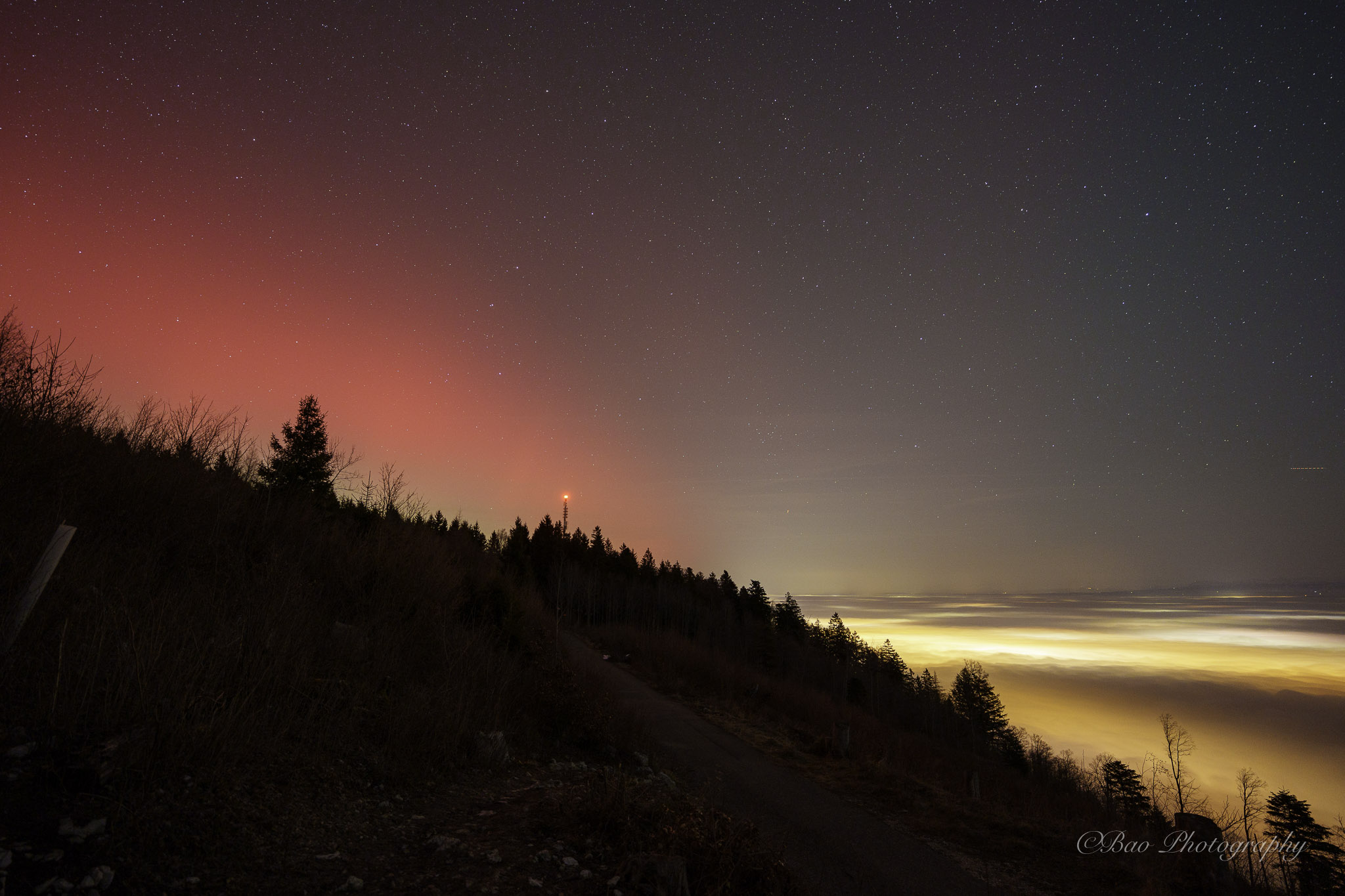 Aurora borealis with a red and pink glow over the Magglingen ridge silhouette with a sea of fog below lit by city lights and a starry sky above, with a telecommunications tower visible on the ridgeline