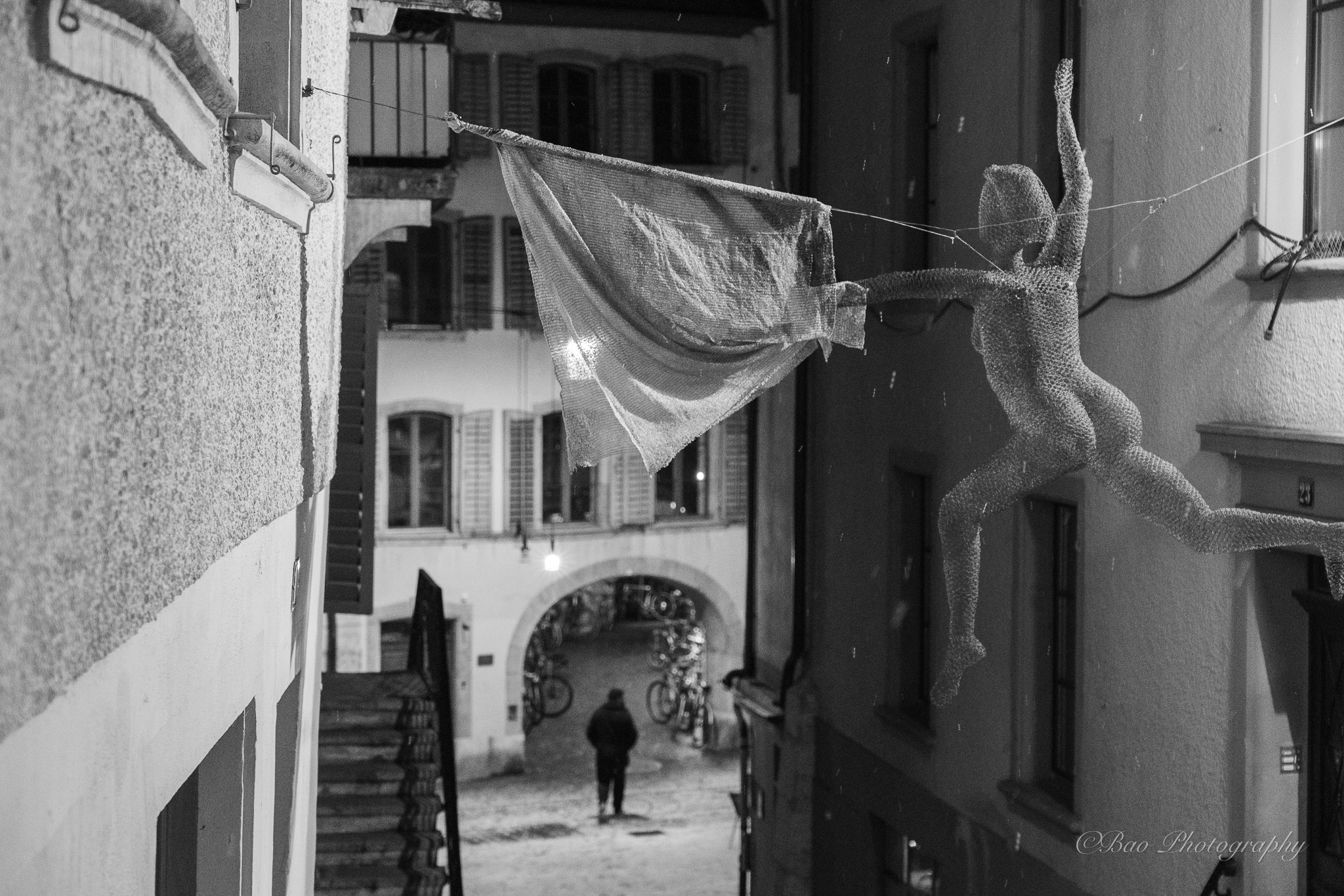 Black and white photograph of a wire mesh figure with a cape suspended between buildings in the old town of Biel, with a person walking through an arched passage below and snowflakes falling